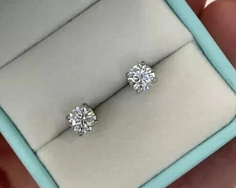 May include: A pair of diamond stud earrings presented in a light blue jewellery box. The earrings are round, with multiple facets. The box has a white interior and is held by a person.