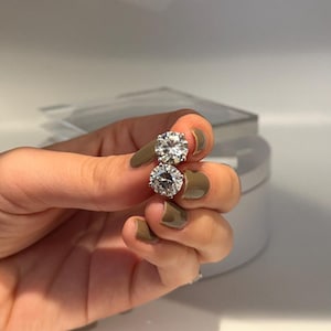 May include: A pair of sparkling, round-cut diamond stud earrings. The earrings are held up against a neutral background, showcasing their brilliance. The diamonds are set in a classic, prong setting, reflecting light and adding a touch of elegance.