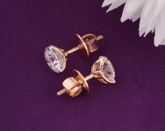 May include: A pair of gold stud earrings with clear, round-cut gemstones. The earrings are set on a purple background, with a white flower in the upper right corner. The earrings have a screw-back closure.