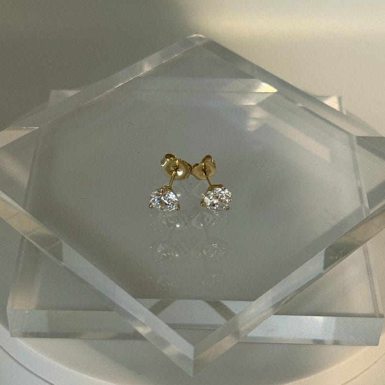 May include: A pair of gold stud earrings with round, clear gemstones. The earrings are displayed on a clear, geometric stand, reflecting light. The setting appears to be a classic four-prong design.