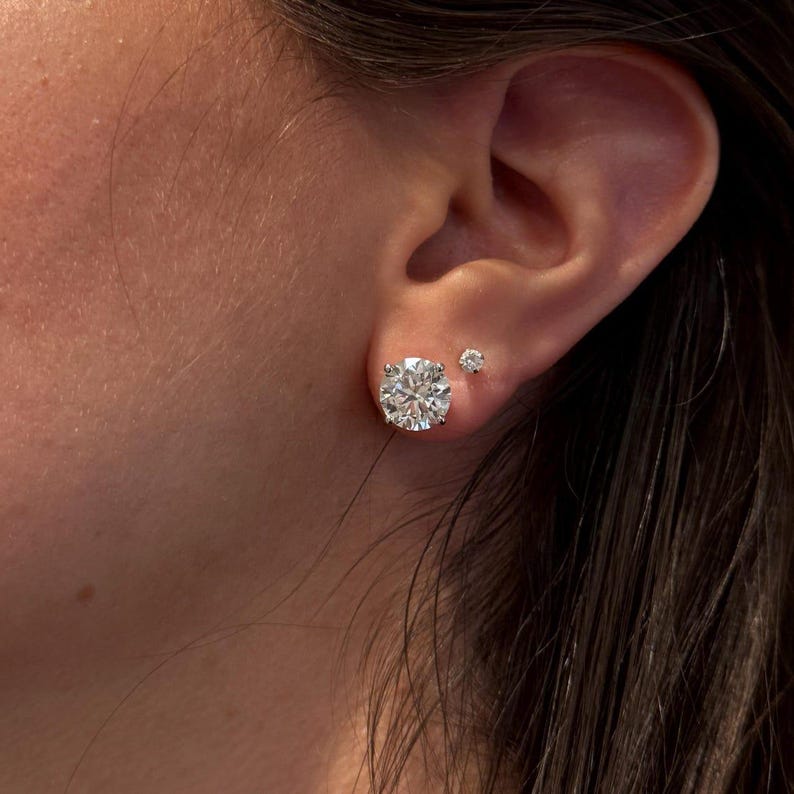May include: A close-up of a person's ear with two diamond stud earrings. The larger earring is a round-cut diamond, and the smaller earring is a smaller round diamond. The earrings sparkle and reflect light.