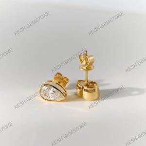 May include: A pair of gold-colored stud earrings. Each earring features a pear-shaped, clear gemstone set in a gold bezel. The earrings are displayed on a white surface, showcasing their design and craftsmanship.