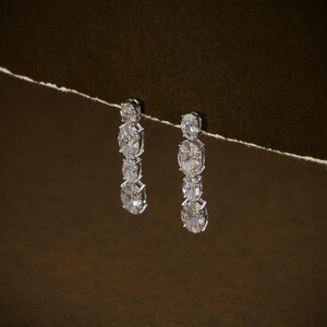 May include: A pair of elegant drop earrings featuring three oval-cut diamonds each, set in a silver-toned metal. The diamonds sparkle against a dark brown background, creating a luxurious and sophisticated look.