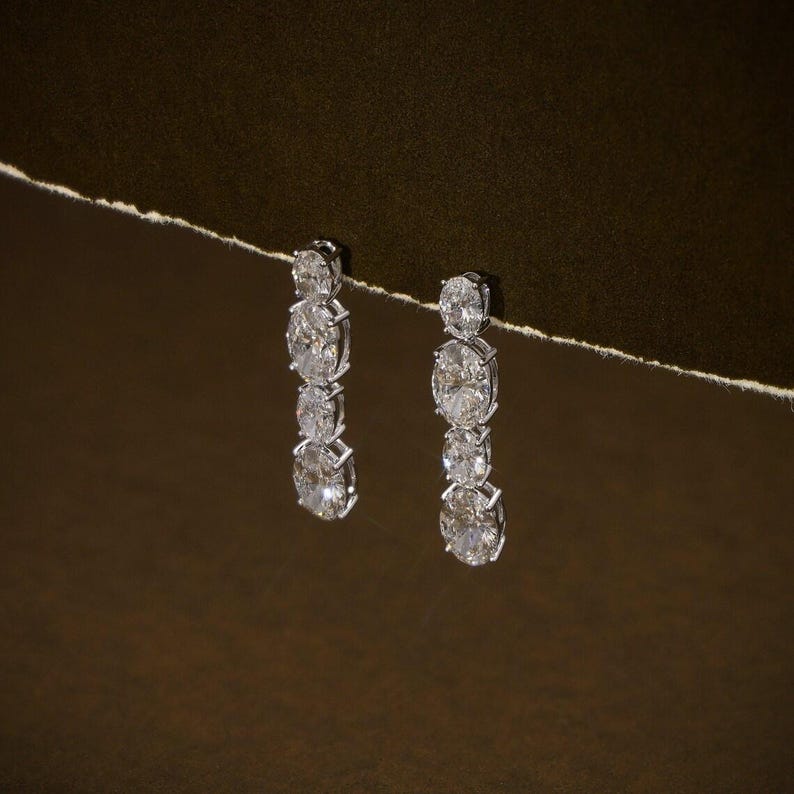 May include: A pair of elegant drop earrings featuring three oval-cut diamonds each, set in a silver-toned metal. The diamonds sparkle against a dark brown background, creating a luxurious and sophisticated look.
