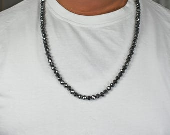May include: A long, beaded necklace with faceted, dark gray beads. The necklace is draped over a white t-shirt, showcasing the jewelry's length and color. The beads appear to be evenly sized and strung together.