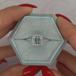 Könnte beinhalten: Ein Ring mit einem hellblauen, quadratisch geschliffenen Edelstein, umgeben von einem Halo aus kleineren, klaren Steinen. Der Ring ist in einem silbernen Band gefasst und wird in einer hellblauen, sechseckigen Ringschatulle präsentiert.