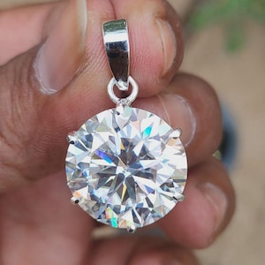 May include: A round, clear gemstone pendant with a silver bail. The gemstone is faceted, reflecting light with a brilliant sparkle. The pendant is held by a hand, showcasing the jewelry's design and clarity.