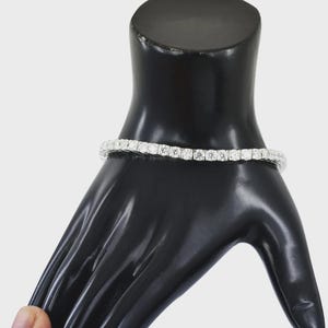 May include: A tennis bracelet with multiple round-cut, clear gemstones set in a row. The bracelet is displayed on a black mannequin hand. The gemstones appear to be of similar size and are closely set together.