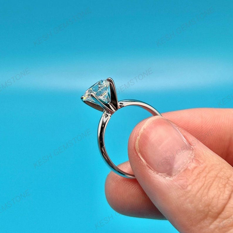 Op de afbeelding: Een zilveren ring met een grote, heldere edelsteen. De ring wordt tegen een helderblauwe achtergrond gehouden, waarbij de sprankelende steen en het gepolijste metaal van de ring worden benadrukt. De edelsteen is hoog geplaatst.