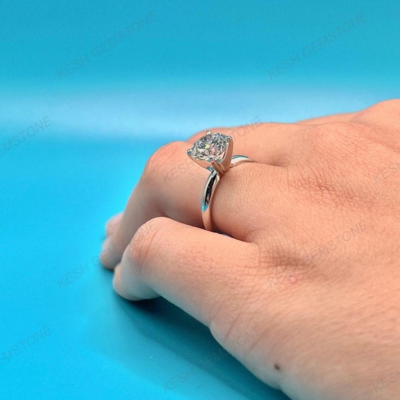 Op de afbeelding: Een zilveren verlovingsring met een grote, ronde, heldere edelsteen. De ring is gezet op een eenvoudige band en wordt op een hand getoond tegen een blauwe achtergrond. De edelsteen schittert.