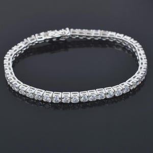 May include: A tennis bracelet with a silver-colored metal setting. The bracelet is adorned with numerous round, clear gemstones, creating a continuous line of sparkle. The clasp is visible, securing the bracelet.