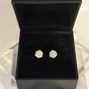 May include: A pair of sparkling diamond stud earrings displayed in an open black jewelry box. The earrings are round, with a brilliant cut, and set in a gold or yellow metal. The box has a velvet-like interior.
