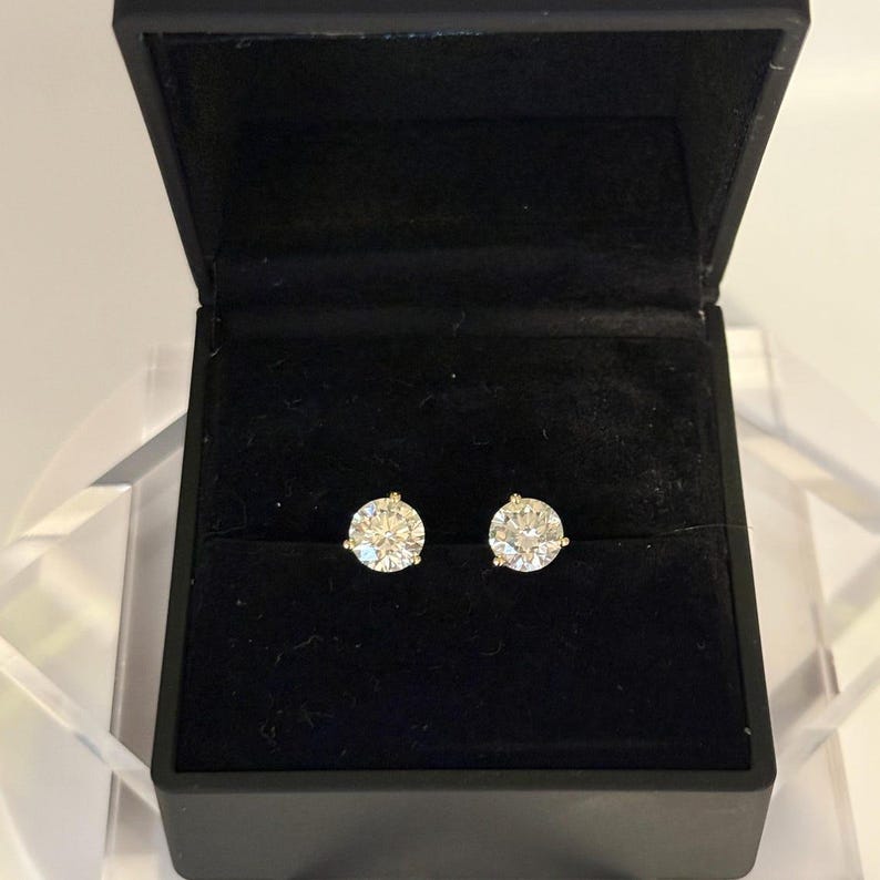 May include: A pair of sparkling diamond stud earrings displayed in an open black jewelry box. The earrings are round, with a brilliant cut, and set in a gold or yellow metal. The box has a velvet-like interior.