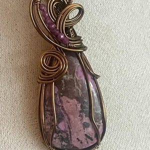 Peut inclure: Pendentif artisanal avec une pierre en forme de goutte d'eau aux nuances de violet et de marron, sertie dans du fil de cuivre. Le travail du fil comprend des volutes décoratives et des accents de petites perles violettes. Le pendentif est suspendu à un cordon noir.