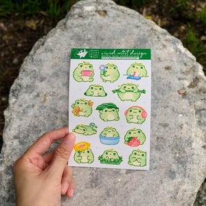 May include: A sticker sheet featuring various green frog illustrations with different accessories and activities. The sheet includes the text "vivid mist design". The stickers are held by a hand against a gray rock background.