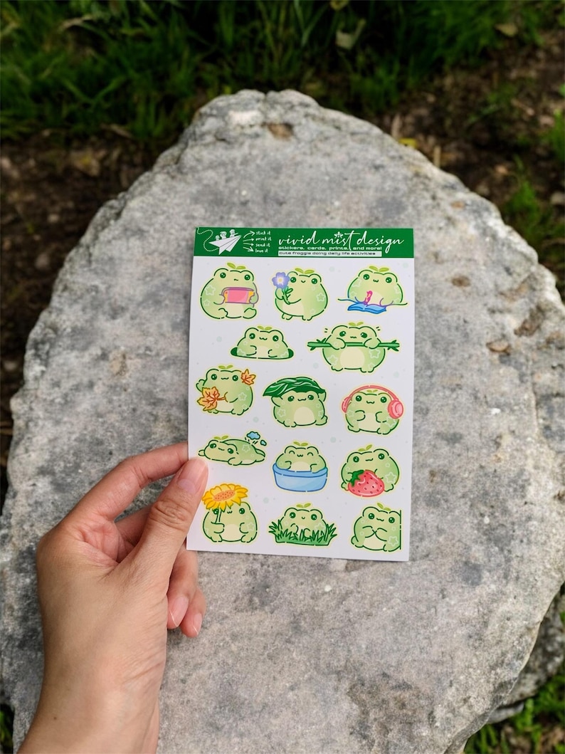 May include: A sticker sheet featuring various green frog illustrations with different accessories and activities. The sheet includes the text "vivid mist design". The stickers are held by a hand against a gray rock background.