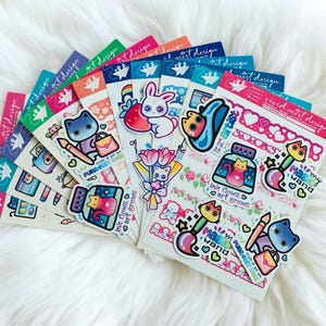 May include: A collection of colorful sticker sheets with various designs, including cats, bunnies, paintbrushes, and floral elements. The stickers feature text like "PURR-FECT PACK" and "ink flows, art grows." The sheets are arranged on a fluffy white surface.