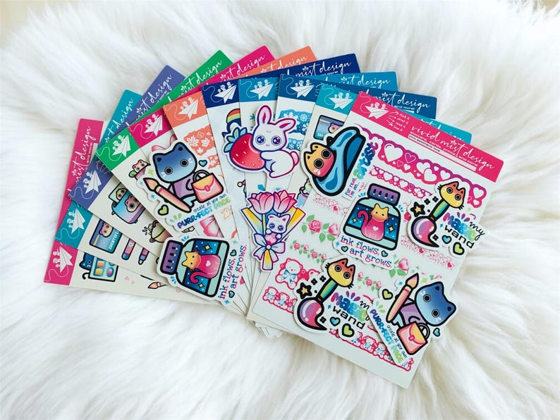 May include: A collection of colorful sticker sheets with various designs, including cats, bunnies, paintbrushes, and floral elements. The stickers feature text like "PURR-FECT PACK" and "ink flows, art grows." The sheets are arranged on a fluffy white surface.