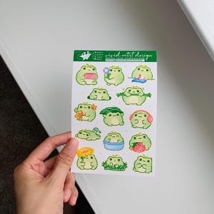 Other frogs are shown enjoying simple pleasures such as listening to music with pink headphones, holding a strawberry nearly as big as itself, resting with sleepy eyes, or clutching a long green branch with a content grin. The frogs share a consistent color palette of soft green tones, creamy bellies, rosy cheeks, and tiny star accents that add charm without overwhelming the design.