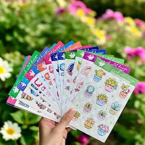 May include: A hand holds a fan of colorful sticker sheets with various designs. The stickers feature illustrations of succulents, floral patterns, and other cute graphics. The text "vivid mist design" is printed on the top right corner of each sheet.
