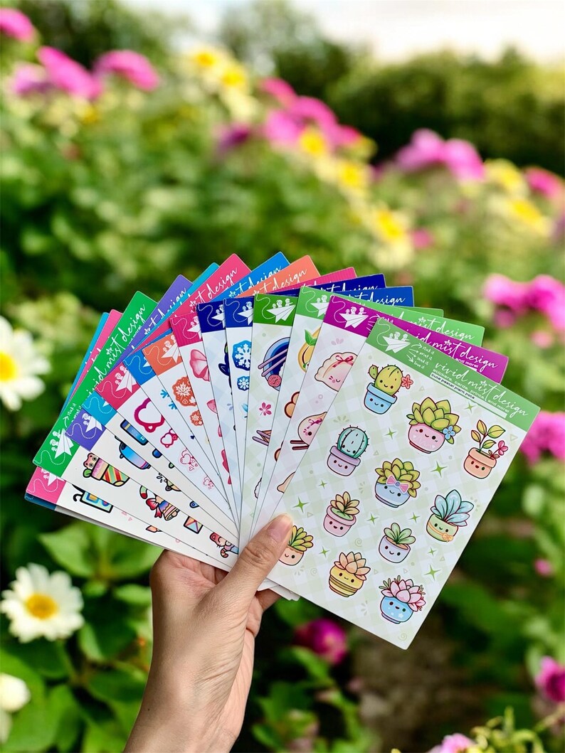 May include: A hand holds a fan of colorful sticker sheets with various designs. The stickers feature illustrations of succulents, floral patterns, and other cute graphics. The text "vivid mist design" is printed on the top right corner of each sheet.