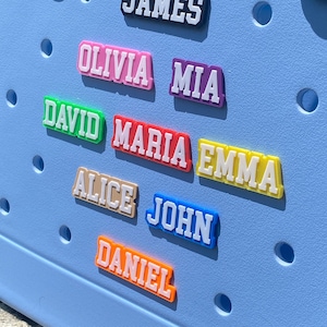 May include: A selection of brightly coloured name tags, each bearing a different name in white block lettering. The tags are in various colours, including black, pink, purple, green, red, yellow, beige, blue, and orange. They are attached to a light blue surface.