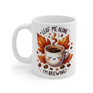 Puede incluir: Taza de café de cerámica blanca con un diseño de taza de café de dibujos animados. La taza presenta el texto "LEAF ME ALONE I'M BREWING!" con hojas de otoño y granos de café. Un diseño encantador y acogedor.