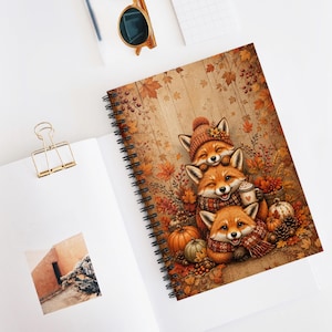 May include: A spiral-bound notebook featuring an illustration of three stacked foxes wearing autumn-themed scarves and hats, surrounded by fall foliage, pumpkins, and pinecones. The cover art displays a warm, rustic color palette with orange and brown tones.