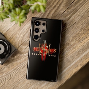 May include: Black phone case with the words "JESUS SAVED MY LIFE" in white and gold lettering. The design includes three nails and a red cross. The case is on a wooden surface.