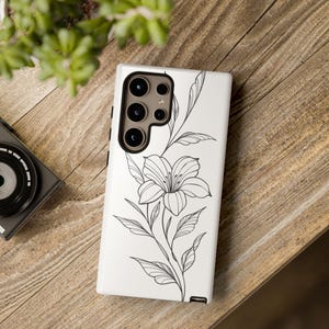 May include: White phone case featuring a black floral line art design. The design includes a large flower with detailed petals and leaves. The phone case is on a wooden surface, with a camera and greenery in the background.