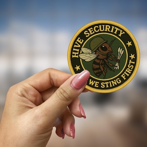 May include: A yellow and green embroidered patch with a bee wearing a helmet and holding a stinger. The text on the patch reads "Hive Security We Sting First".