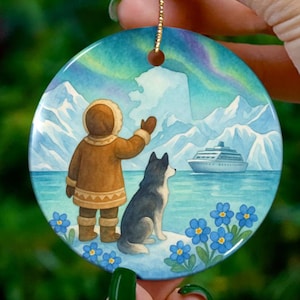 May include: Round ceramic ornament featuring a watercolor scene of a person in a brown parka with a husky dog, mountains, and a cruise ship. The background shows an aurora borealis. Blue flowers are in the foreground. A gold string is attached for hanging.