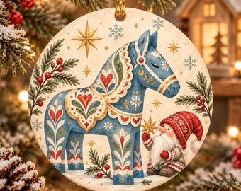 May include: A round ceramic Christmas ornament featuring a blue Dala horse with intricate red, green, and white floral patterns. A small gnome with a red hat and white beard stands beside the horse, holding a gold star. The background includes snowflakes and pine branches.