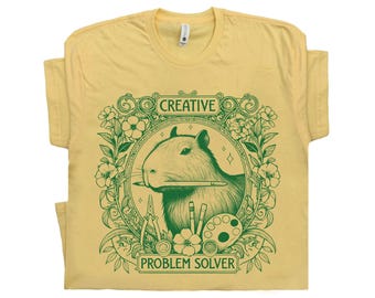 May include: Yellow t-shirt with a green illustration of a capybara holding a paintbrush in its mouth. The illustration is surrounded by flowers and the text "CREATIVE PROBLEM SOLVER".