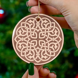 May include: A round, brown ornament with a gold string for hanging. The ornament features a detailed, symmetrical, light brown Celtic knot design. The background is blurred, suggesting a festive setting.