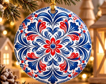 May include: A round ceramic Christmas ornament featuring a symmetrical folk-art pattern in vibrant red and blue on a white background. The design includes stylized floral motifs, swirling leaves, and small red dots, suspended by a gold cord.