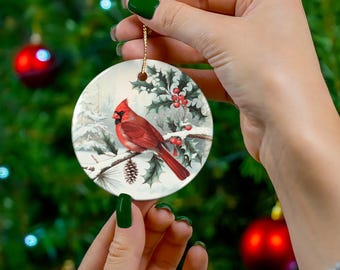 May include: A round white ceramic ornament featuring a detailed illustration of a bright red cardinal bird perched on a snow-covered pine branch with green holly leaves and red berries, suspended by a thin gold cord against a blurred background.