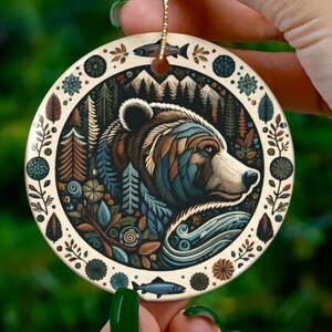 May include: A round ceramic ornament featuring a detailed illustration of a bear's profile, set against a forest and mountain backdrop. The design incorporates shades of brown, blue, and green, with floral and fish motifs around the edge. A gold string is attached for hanging.