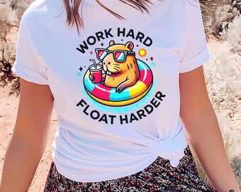 May include: White t-shirt with a cartoon capybara wearing sunglasses and a floatie, holding a drink. The text "Work Hard Float Harder" is printed on the shirt.