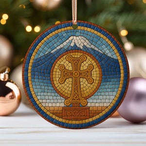 May include: Round Christmas ornament with a mosaic design. The ornament features a cross in the center, surrounded by a blue and yellow mosaic pattern. A mountain is depicted at the top. The ornament is hanging from a gold ribbon.