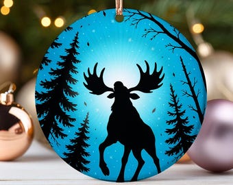 May include: A round Christmas ornament with a blue background featuring a black silhouette of a moose with large antlers. The moose is standing in a forest with trees and falling leaves. The ornament is hanging from a gold ribbon.