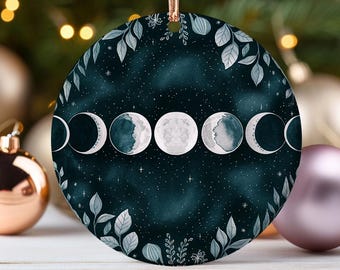 May include: Round Christmas ornament featuring a watercolor design of the phases of the moon against a dark teal starry background. The ornament is trimmed with a border of gray leaves and foliage, and has a gold ribbon for hanging.