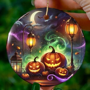 May include: Round Halloween ornament featuring a spooky night scene with glowing jack-o'-lanterns, street lamps, and bats under a crescent moon. The ornament has a gold string for hanging and is set against a blurred green background.