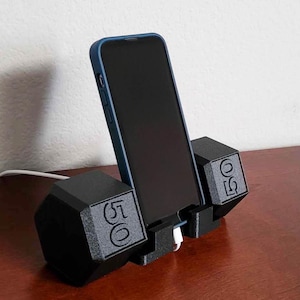 May include: A black phone stand shaped like a dumbbell with the text "50" on the side. The stand is holding a black smartphone.