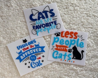 May include: Three white signs with blue, orange, and red lettering. The signs feature cat-themed sayings: "Cats are my favorite people", "Visitors must be approved by the cat", and "Less people more cats..."
