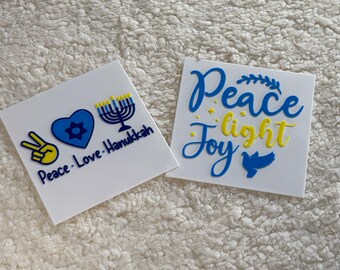 May include: Two white cards with blue and yellow lettering. The first card says "Peace. Love. Hanukkah" with a peace sign, a heart with a Star of David, and a menorah. The second card says "Peace light Joy" with a dove.