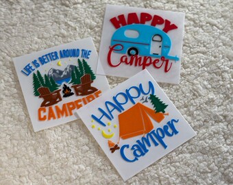 May include: Three white vinyl stickers with camping themes. The first sticker says "Life is better around the campfire". The second sticker says "Happy Camper" with a blue camper van. The third sticker says "Happy Camper" with an orange tent.