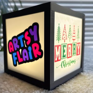 May include: A black wooden box with a light inside. The box has a white background with the words "Arts Flair" in colorful letters on one side and "Merry Christmas" with a row of green Christmas trees on the other side.