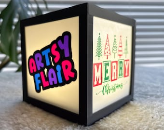 May include: A black wooden box with a light inside. The box has a white background with the words "Arts Flair" in colorful letters on one side and "Merry Christmas" with a row of green Christmas trees on the other side.