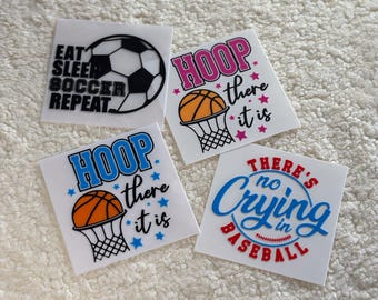 May include: Four white stickers with colorful text and images. The stickers feature the phrases "Eat Sleep Soccer Repeat", "Hoop There It Is", "Hoop There It Is", and "There's No Crying In Baseball".